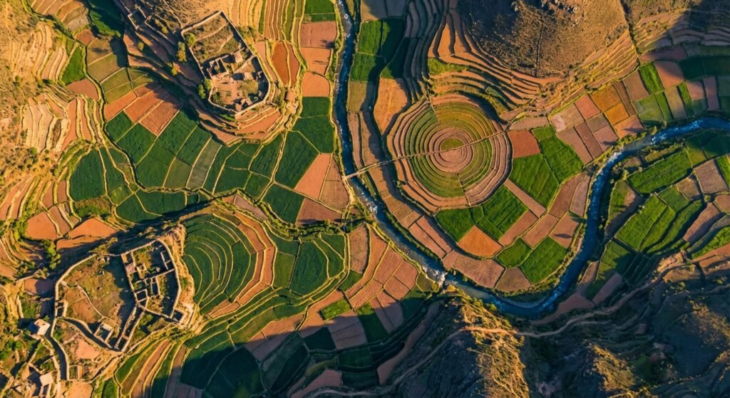 Vista aérea de campos agrícolas aterrazados de colores verdes y rojizos, con un río sinuoso a través de un valle montañoso, estructuras de ruinas antiguas de piedra y un gran campo de terrazas circulares, todo iluminado por la luz del sol.} {